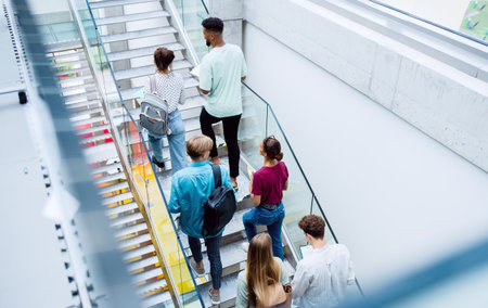 Rear view of university students walking up the stairs indoors, looking at camera and waving.