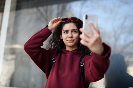 Portrait of young woman with smartphone standing outdoors on street in city, taking selfie.