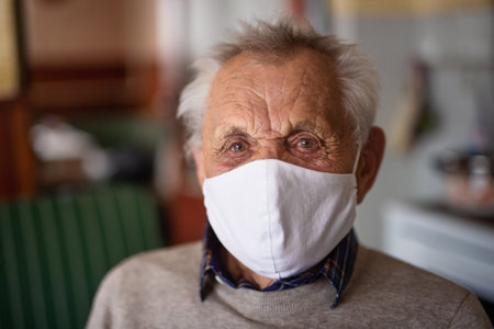 Portrait of elderly man with face mask looking at camera indoors at home, coronavirus concept.の写真素材