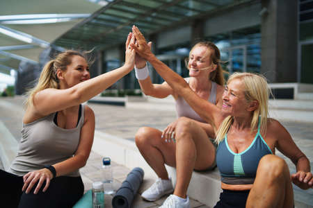 Group of young and old women sitting after exercise outdoors in city, giving high five.の写真素材