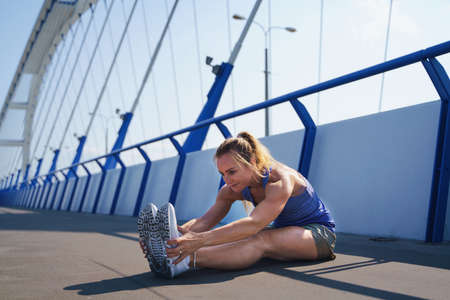 Mid adult woman runner stretching outdoors on bridge in city, healthy lifestyle concept.