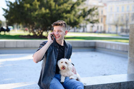 Portrait of young man with dog outdoors in city, using smartphone.の写真素材