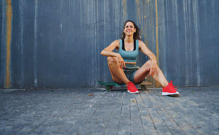 Front view portrait of mid adult woman sitting on skateboard outdoors in city, resting.の写真素材