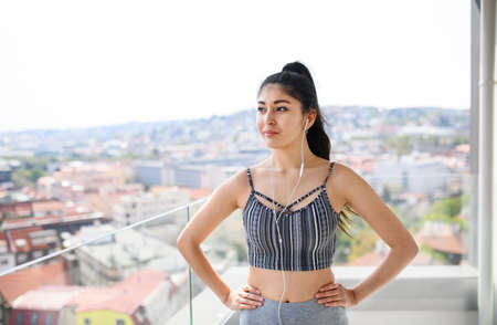 Portrait of young sport woman with earphones standing on balcony outdoors in city, resting.