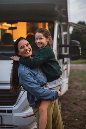 Mother with daughter standing by car outdoors in campsite at dusk, caravan family holiday trip.の写真素材