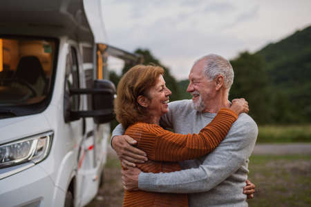 Senior couple standing and hugging outdoors at dusk, caravan holiday trip.の写真素材