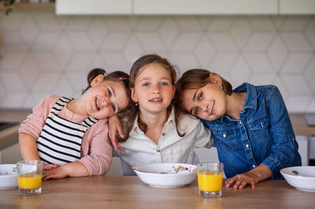 Portrait of three girls sisters indoors at home, looking at camera when eating breakfast.