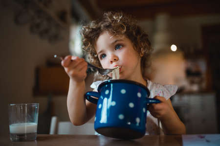 Sad small girl sitting at table indoors at home, eating spaghetti.