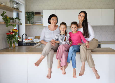 Happy small girls with mother and grandmother sitting on kitchen counter indoors at home, looking at camera.の写真素材