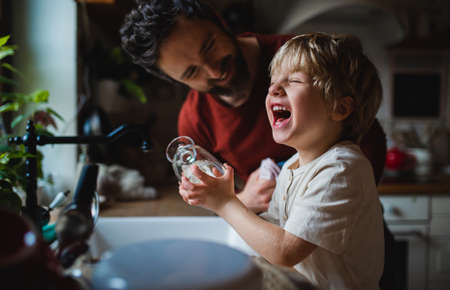 Small laughing boy with father having fun when washing dishes indoors at home, daily chores concept.の写真素材
