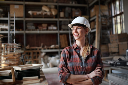 Portrait of mid-adult female carpenter standing in carpentery workshop, looking aside and smiling. Small business concept.の写真素材