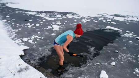 Active senior woman in swimsuit breaking ice with axe outdoors in winter, cold therapy concept.の写真素材