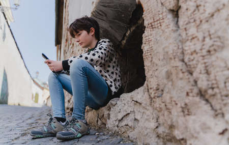 Low angle view of preteen girl with smartphone sitting outdoors in town.