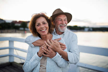 Happy senior couple hugging outdoors on pier by sea, looking at camera.