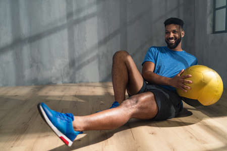 Young African American sportsman sitting and lifting a medicine ball indoors, workout training concept.の写真素材