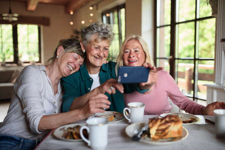 Happy senior friends having coffee and cake and taking selfie indoors at home.の写真素材