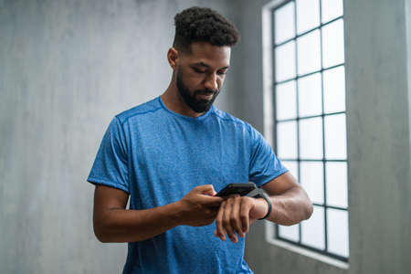 Happy young African American sportsman standing indoors at gym, using smartphone and smartwatch.の写真素材