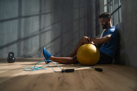 Young African American sportsman sitting and using smartwatch indoors in gym, workout training concept.の写真素材