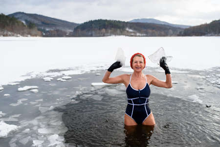 Front view of active senior woman in swimsuit stnading in water hole in frozen lake holding ice, looking at camera outdoors in winter, cold therapy concept.の写真素材