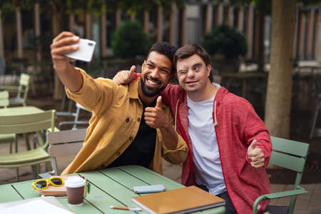 Young man with Down syndrome and his mentoring friend sitting and taking selfie outdoors in cafeの写真素材