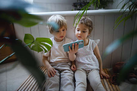 Cute small children sitting on floor indoors at bathroom, using smartphone.