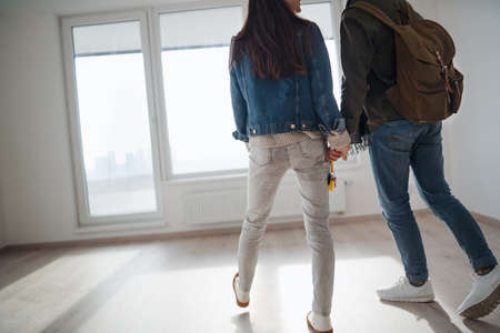 Rear view of unrecognizable young couple owners holding hands in new flat, moving in.