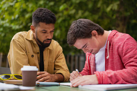 Young man with Down syndrome with his mentoring friend sitting outdoors in cafe and studying.の写真素材