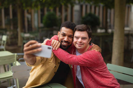 Young man with Down syndrome and his mentoring friend sitting and taking selfie outdoors in cafeの写真素材