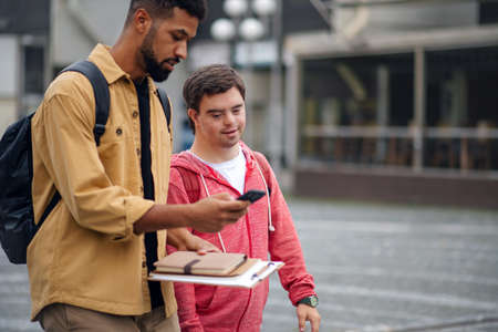 Young man with Down syndrome with his mentoring friend with smartphone walking and talking outdoorsの写真素材