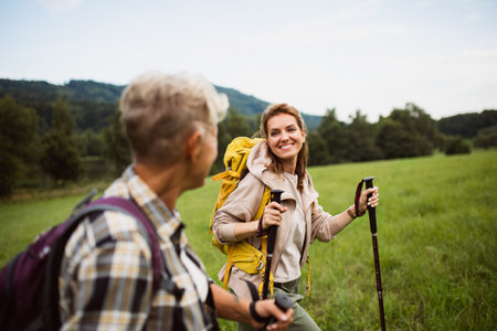 Happy mid adult woman with trekking poles hiking with active senior mother outdoors in nature.