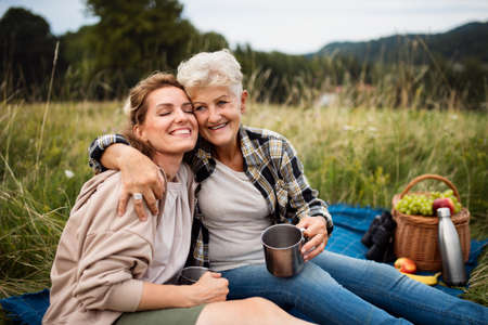 Happy senior mother embracing adult daughter when sitting and having picnic outdoors in nature.の写真素材