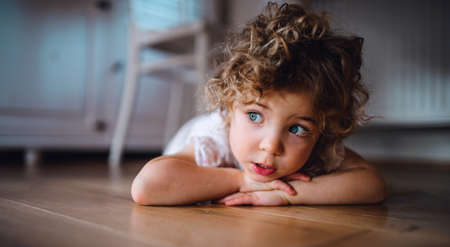 Portrait of cute small girl lying on floor under table indoors at home, looking away.の写真素材