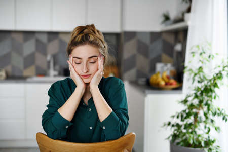 Portrait of tired young woman student sitting and relaxing with eyes closed indoors in kitchen.