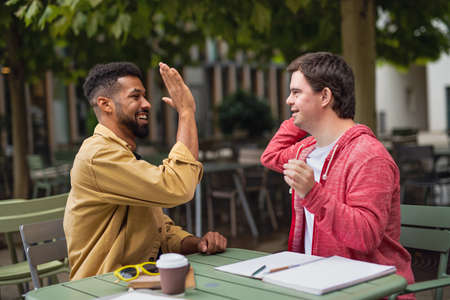 Young man with Down syndrome with mentoring friend sitting outdoors in cafe celebrating success.