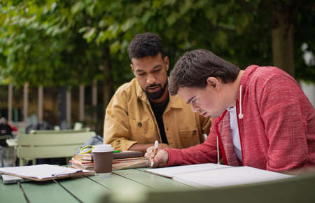 Young man with Down syndrome with his mentoring friend sitting outdoors in cafe and studying.の写真素材