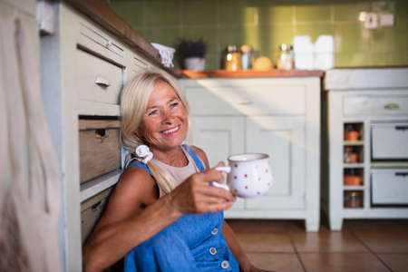 Happy senior woman sitting on floor and drinking tea indoors in kitchen.