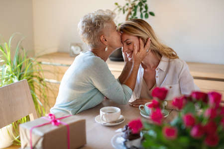 Happy senior mother having coffee with adult daughter indoors at home, kissing her on forehead.の写真素材