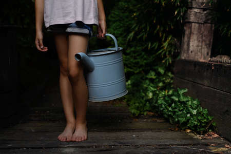 Low section of little girl in shorts holding watering can outdoors in garden.の写真素材