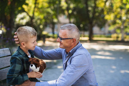 Senior man squatting and talking to grandson outdoors on bench in park.の写真素材