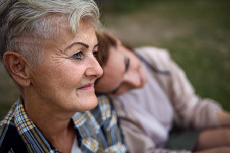 Close up of senior mother sitting on grass with adult daughter leaning her head on her outdoors.の写真素材