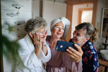 Happy senior women friends in bathrobes taking selfie indoors at home, selfcare concept.の写真素材