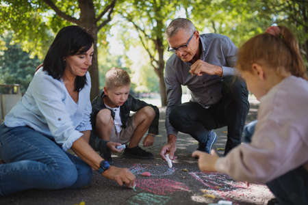 Senior couple with grandchildren drawing with chalks on pavement outdoors in park.
