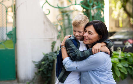 Happy schoolboy hugging his grandmother waiting for him after school outdoors in street.