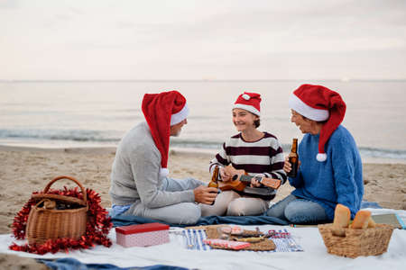 Happy senior couple with granddaughter sitting on blanket and having picnic outdoors on beach by sea.の写真素材