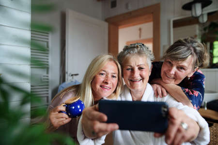 Happy senior women friends in bathrobes taking selfie indoors at home, selfcare concept.の写真素材