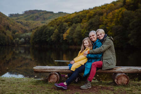 Small girl with mother and grandmother sitting on bench and looking at camera outoors by lake.の写真素材