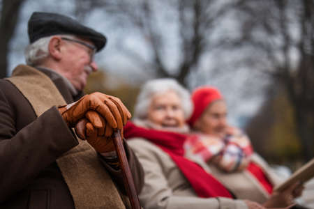 Group of happy senior friends sitting on bench in town park in autumn, focus on mans hands in glovesの写真素材