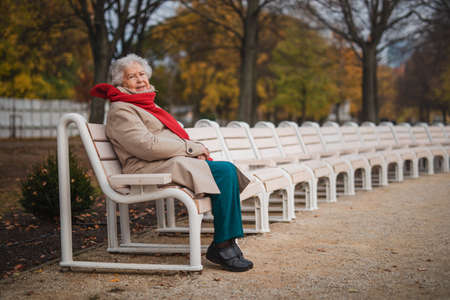 Senior woman sitting on bench in town park in autumn, looking at camera.の写真素材