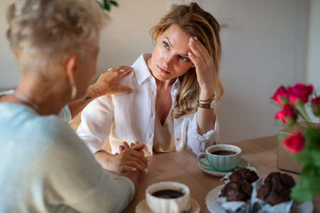 Senior mother consoling sad adult daughter indoors at home, sitting and having coffee.の写真素材