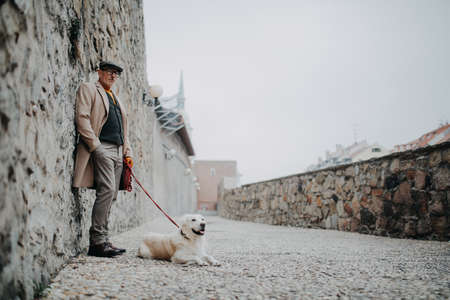 Senior man looking at camera and leaning against the wall when walking his dog outdoors in city.の写真素材
